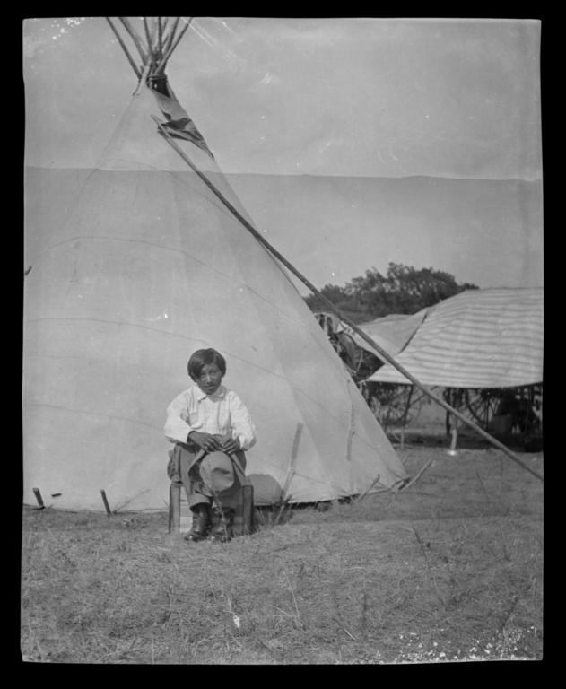 Native American Boy With a Hat Sitting in front of a Tipi